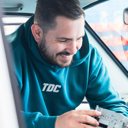 Man in a teal hoodie with 'TDC' logo holding a device inside a vehicle.