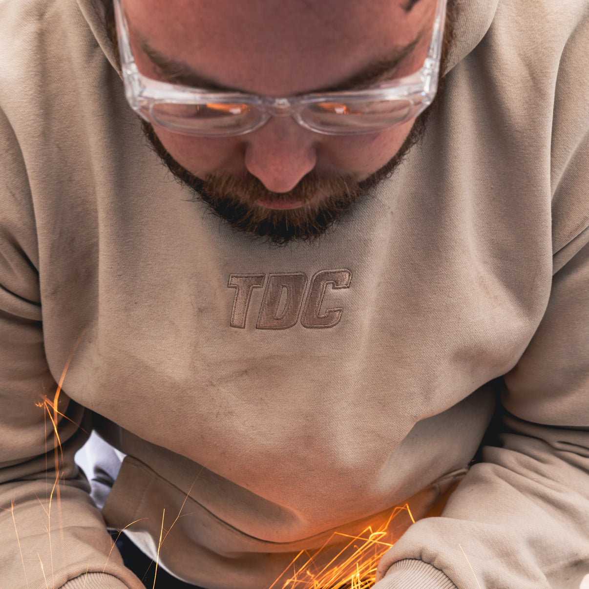 Person wearing a beige hoodie with 'TDC' logo, using a red tool with sparks flying.