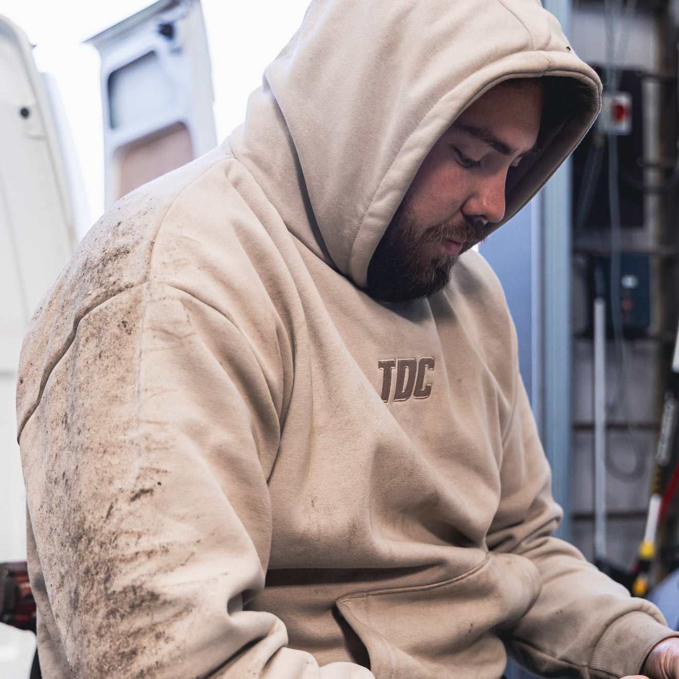 Person wearing a beige hoodie with 'TDC' logo, sitting and working on a laptop in an indoor setting.